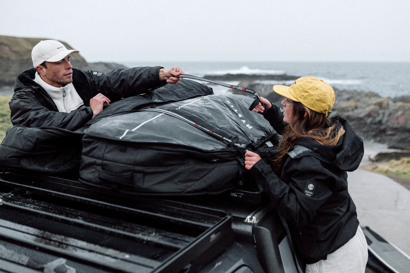 Two surfers strapping a large black board bag onto a car roof rack beside the sea