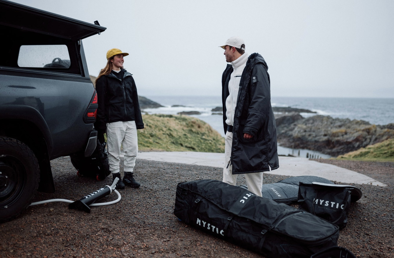 Two surfers in changing robes beside a 4x4 on the coast with Mystic board bags on the ground
