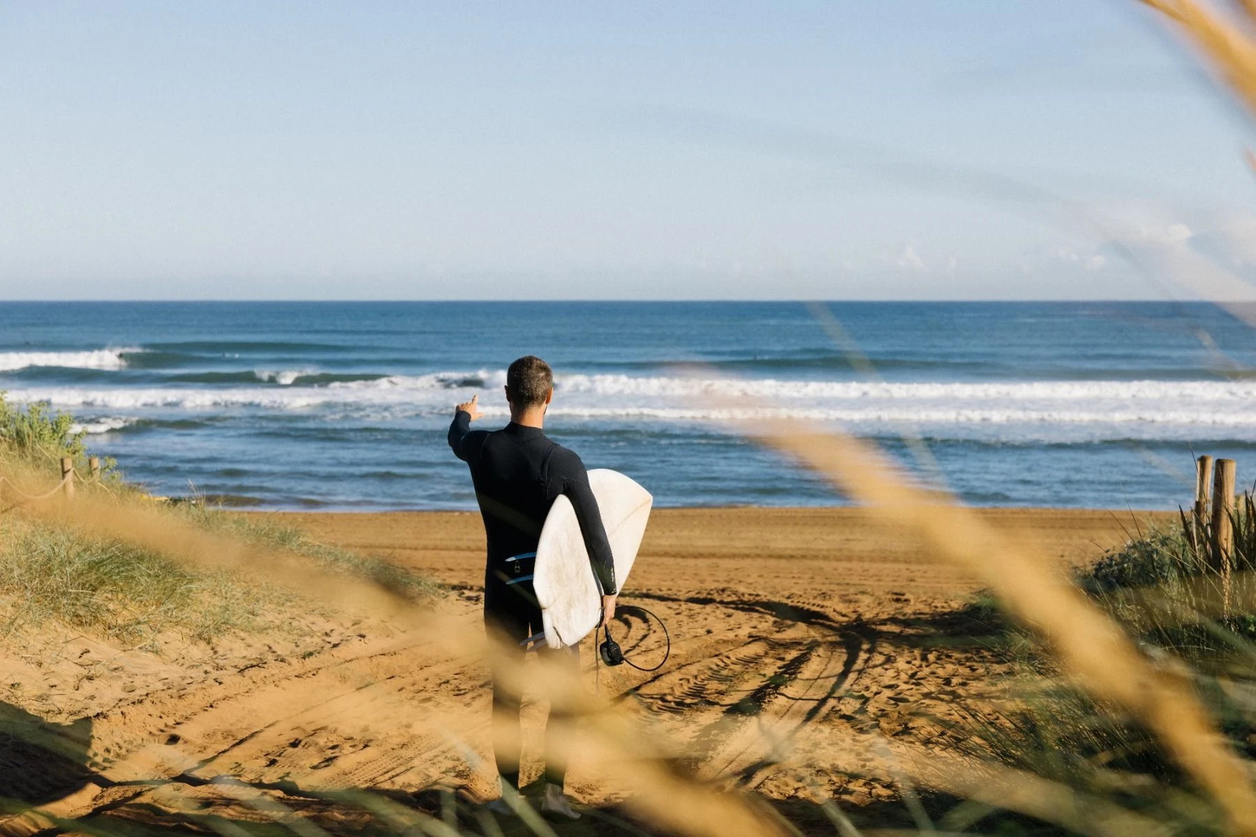 Surfeur en combinaison noire descendant un sentier de dune sablonneux vers la plage, planche sous le bras