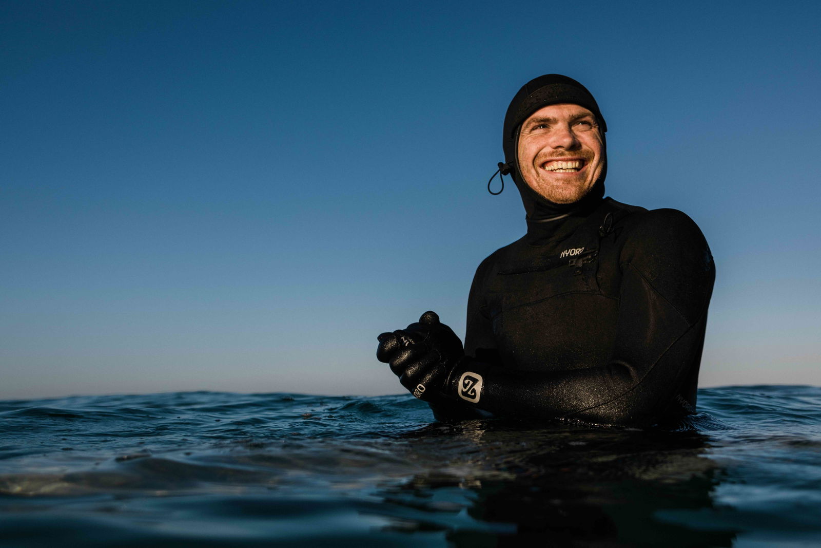 Homme souriant en combinaison noire à cagoule et gants, immergé jusqu'à la poitrine dans la mer