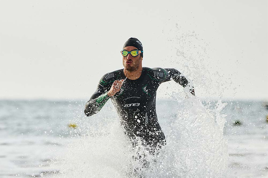 Homme en combinaison de nage ZONE3 avançant dans les vagues, entouré d'embruns