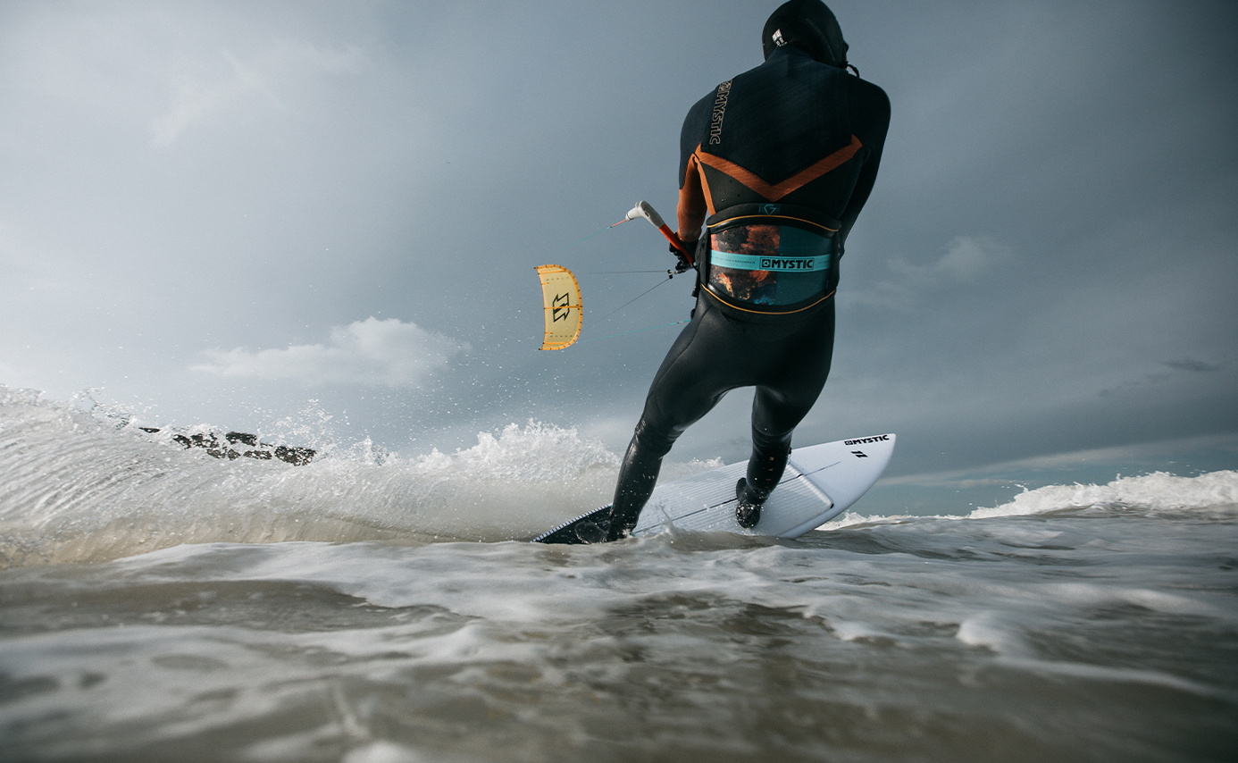 Kitesurfeur en combinaison noire à cagoule taillant une vague déferlante sur un surf, aile jaune au-dessus